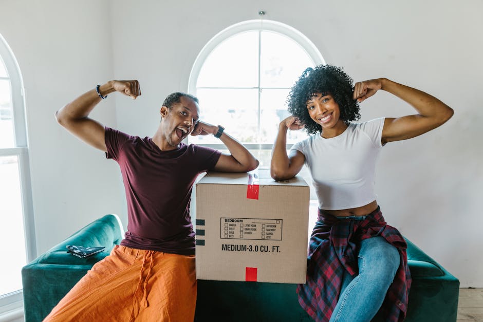 A man and woman inside a bright room with white walls and a large arched window in the background, are smiling and flexing their arms in a playful manner while sitting on a green sofa. Between them, t